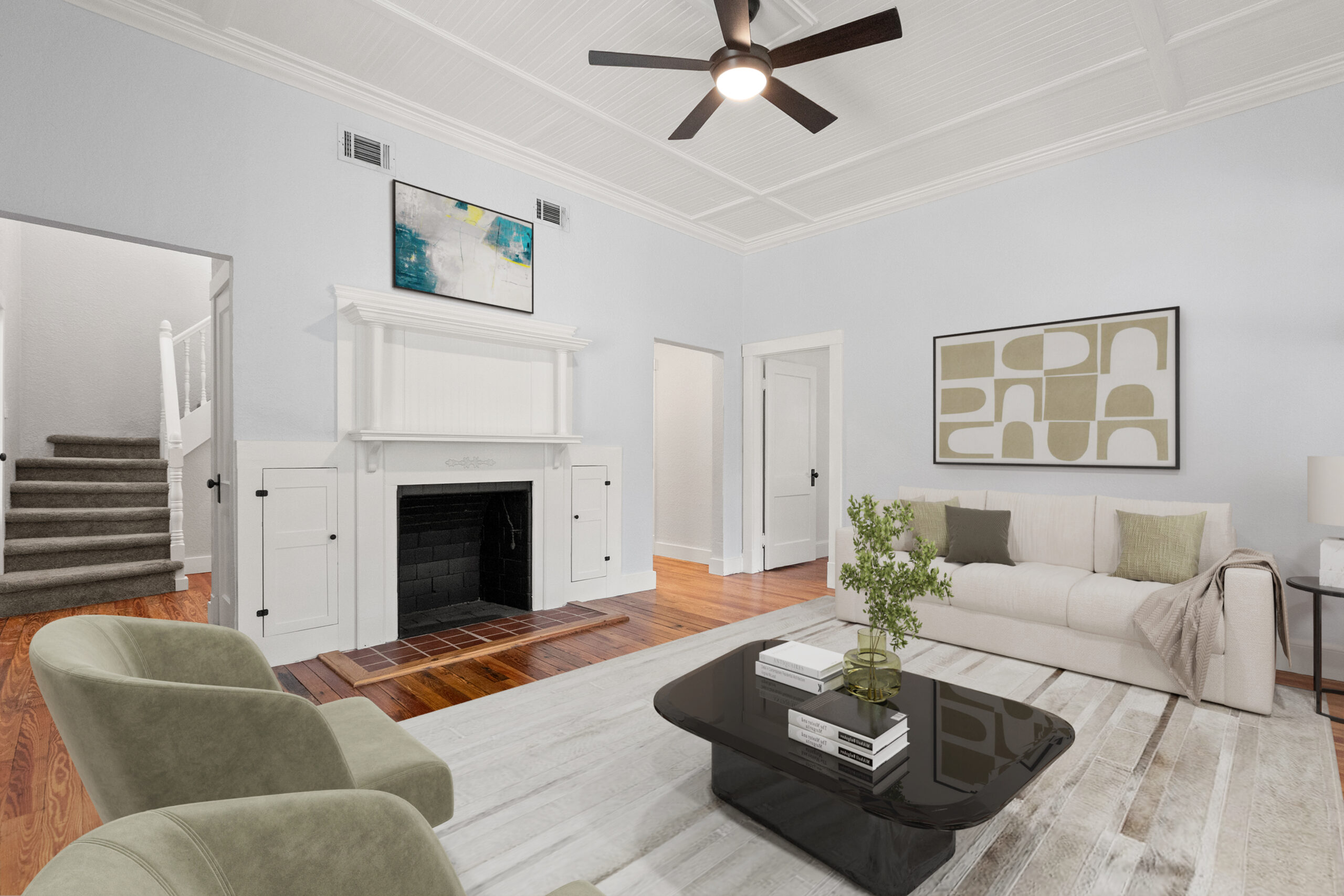 Modern living room with a neutral color palette, featuring a white sofa, green accent chairs, a black coffee table with books, and a decorative plant; includes a fireplace, artwork on the wall, and a staircase in the background, showcasing inviting real estate photography.